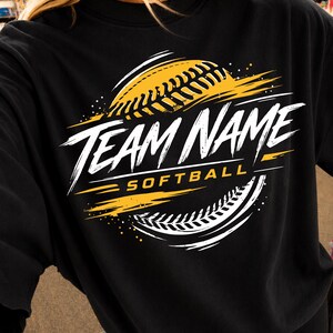 May include: A black long-sleeve shirt featuring a graphic design with a yellow and white softball, brushstroke accents, and the text "TEAM NAME" in a bold white font, with the word "SOFTBALL" printed in yellow underneath the main text.