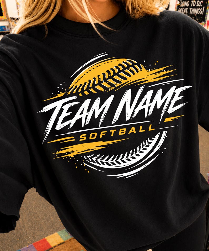 May include: A black long-sleeve shirt featuring a graphic design with a yellow and white softball, brushstroke accents, and the text "TEAM NAME" in a bold white font, with the word "SOFTBALL" printed in yellow underneath the main text.