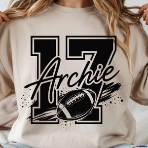 May include: A beige crewneck sweatshirt featuring a large black graphic print of the number 17 with the name Archie written in a cursive script across the center, accompanied by a black and white football illustration with motion lines.