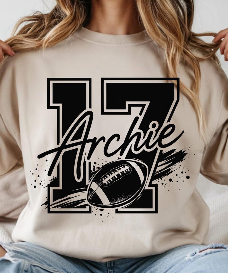 May include: A beige crewneck sweatshirt featuring a large black graphic print of the number 17 with the name Archie written in a cursive script across the center, accompanied by a black and white football illustration with motion lines.