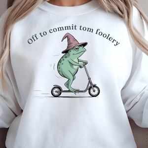 Peut inclure: Sweat-shirt blanc avec l'inscription "Off to commit tom foolery" en arc de cercle. Le motif représente une grenouille verte portant un chapeau de sorcier, sur un scooter. L'illustration est de style cartoon.