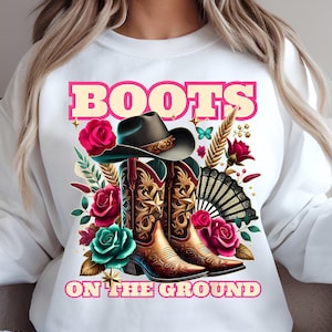 May include: White sweatshirt with the text "BOOTS ON THE GROUND" in pink letters. The design features a pair of brown cowboy boots with a star design, a cowboy hat, and pink and teal flowers.