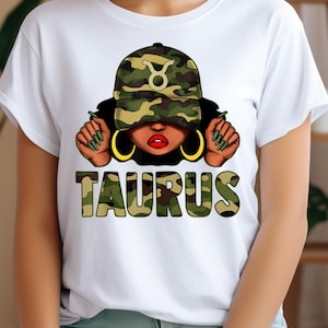 May include: White t-shirt with a graphic of a woman wearing a camouflage hat with a Taurus zodiac symbol. The text "TAURUS" is written in camouflage letters below the graphic.