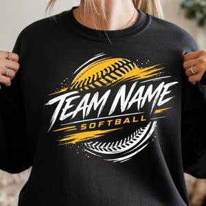 May include: A black crewneck sweatshirt featuring a graphic design of a yellow softball with white stitching. The text "TEAM NAME" is printed in a bold, white, distressed font across the centre, with the word "SOFTBALL" in yellow below it.