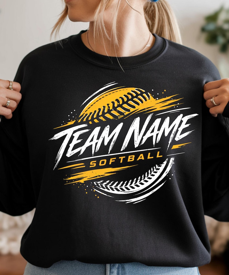 May include: A black crewneck sweatshirt featuring a graphic design of a yellow softball with white stitching. The text "TEAM NAME" is printed in a bold, white, distressed font across the centre, with the word "SOFTBALL" in yellow below it.