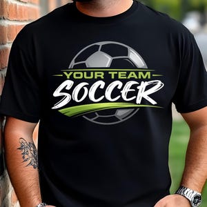 May include: Black t-shirt with a soccer ball graphic. The design includes the text "-YOUR TEAM- SOCCER" in white and green. The soccer ball is gray and white. The shirt is worn by a person.