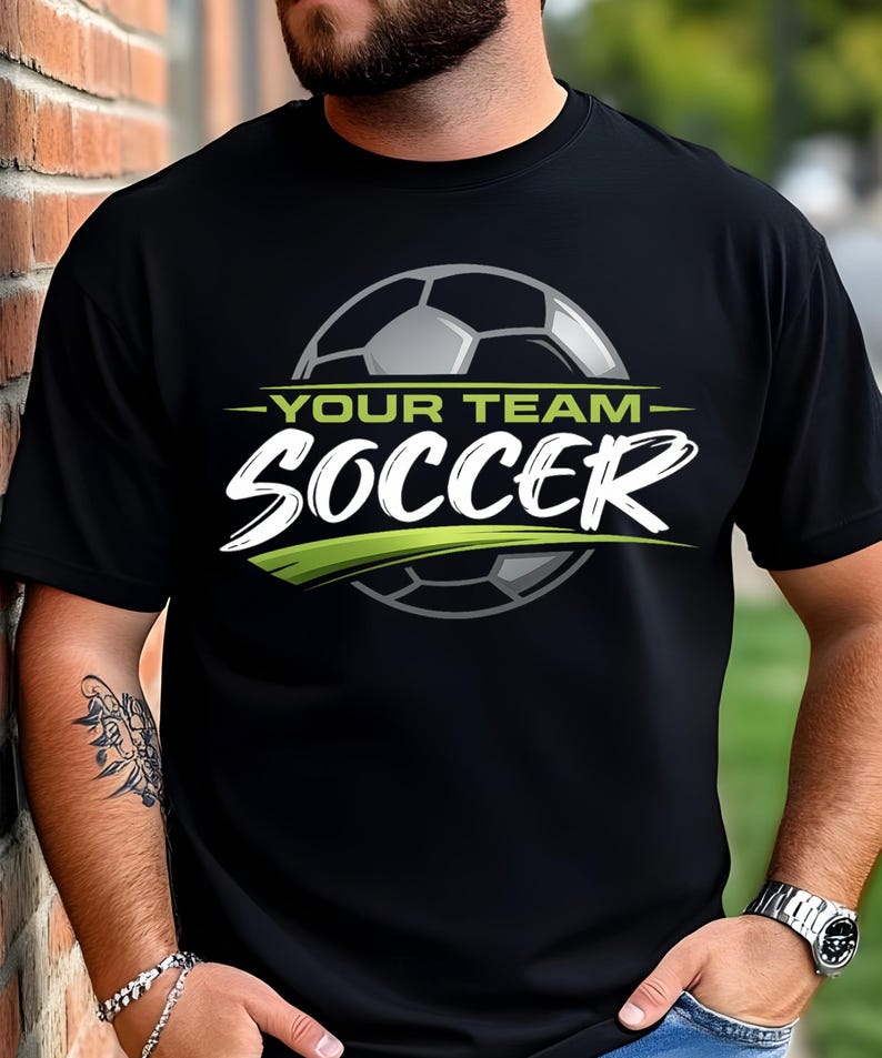 May include: Black t-shirt with a soccer ball graphic. The design includes the text "-YOUR TEAM- SOCCER" in white and green. The soccer ball is gray and white. The shirt is worn by a person.