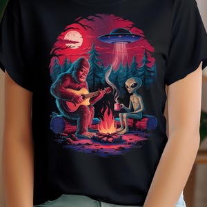 Peut inclure: Un t-shirt noir avec un motif représentant un Bigfoot jouant de la guitare et un extraterrestre buvant dans une tasse près d'un feu de camp. Un OVNI vole au-dessus.