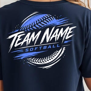 May include: A navy blue t-shirt featuring a graphic on the back with a stylised softball design in white and blue. The graphic includes the text "TEAM NAME" in a bold, brush-stroke font and the word "SOFTBALL" in a clean, sans-serif typeface.