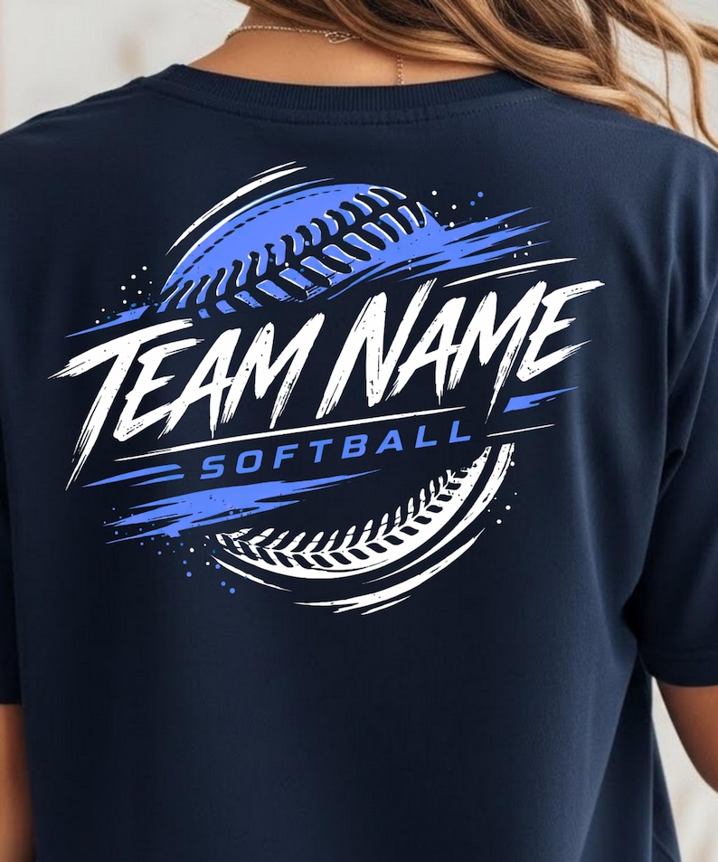 May include: A navy blue t-shirt featuring a graphic on the back with a stylised softball design in white and blue. The graphic includes the text "TEAM NAME" in a bold, brush-stroke font and the word "SOFTBALL" in a clean, sans-serif typeface.