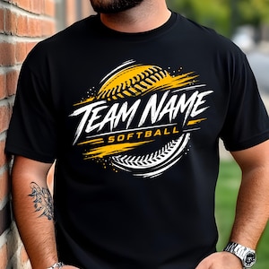 May include: A black short-sleeved t-shirt featuring a graphic design of a softball with yellow and white brushstroke accents. The text "TEAM NAME" is printed in a bold white font, with the word "SOFTBALL" in yellow underneath the main text.