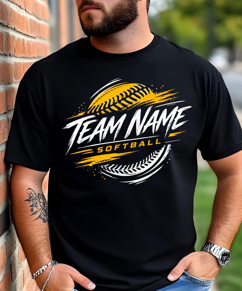 May include: A black short-sleeved t-shirt featuring a graphic design of a softball with yellow and white brushstroke accents. The text "TEAM NAME" is printed in a bold white font, with the word "SOFTBALL" in yellow underneath the main text.