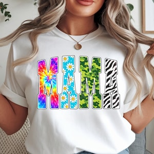 Peut inclure: T-shirt blanc avec le mot "MAMA" en lettres colorées et à motifs. Le "M" est tie-dye, les "A" présentent des motifs de marguerites et le dernier "M" a un imprimé zébré.