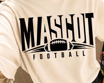 May include: A cream-colored short-sleeve t-shirt featuring a large black graphic print on the chest. The design displays the word MASCOT in bold capital letters above a stylized football icon, with the word FOOTBALL printed in smaller letters below.