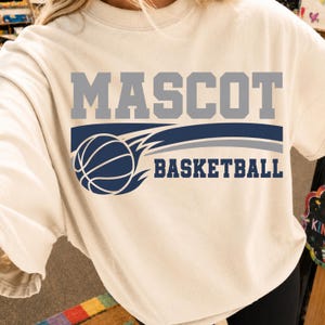 Peut inclure: Un t-shirt à manches courtes de couleur crème avec le mot MASCOT en grandes lettres majuscules grises au-dessus d'un graphique bleu marine représentant un ballon de basket avec des lignes de mouvement et le mot BASKETBALL en bleu marine.