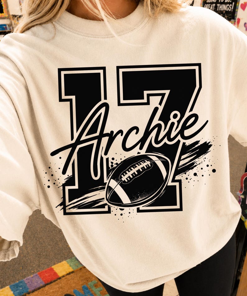 May include: A cream-colored crewneck t-shirt featuring a large black graphic of the number 17 with the name Archie written in a cursive script across the center. A black and white American football illustration is positioned over the number.