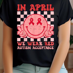 Puede incluir: Camiseta negra con una cara sonriente rosa y roja con signos de paz y cintas rojas sobre los ojos. El texto "IN APRIL WE WEAR RED AUTISM ACCEPTANCE" está impreso en la camiseta.