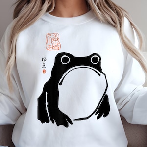 Puede incluir: Sudadera blanca con una ilustración en blanco y negro de una rana y caracteres japoneses en rojo. Los caracteres son "kaeru" (rana) y "o-shimai" (terminado).