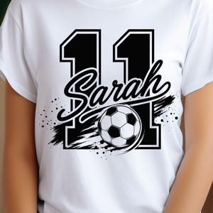 May include: A white short-sleeved t-shirt featuring a large black graphic of the number 11 with the name Sarah written in a cursive script across the center. A black and white soccer ball is positioned over the number, accented by black brush strokes.