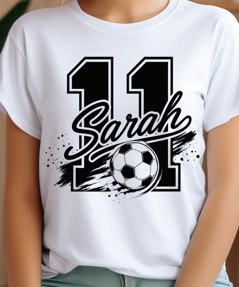 May include: A white short-sleeved t-shirt featuring a large black graphic of the number 11 with the name Sarah written in a cursive script across the center. A black and white soccer ball is positioned over the number, accented by black brush strokes.