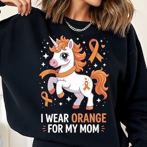 Puede incluir: Sudadera negra con un unicornio blanco, melena y cola naranja, y una cinta naranja. El texto "I WEAR ORANGE FOR MY MOM" está impreso debajo. El diseño incluye estrellas y corazones.