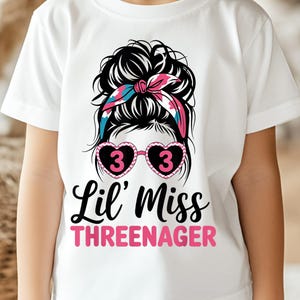 May include: White t-shirt featuring a graphic of a messy bun with a pink and blue headband, heart-shaped sunglasses with the number 3, and the text "Lil' Miss THREENAGER" in black and pink. The shirt is for a child.