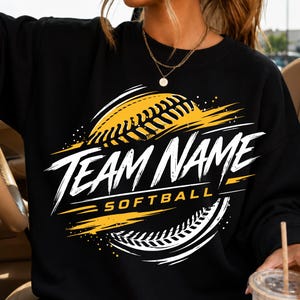 Op de afbeelding: Een zwart sweatshirt met ronde hals en een grafisch ontwerp met een gele softbal en witte penseelstreekaccenten. De tekst TEAM NAME staat in grote witte letters boven het woord SOFTBALL in kleinere gele letters op de borst.