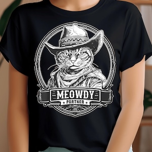 Puede incluir: Camiseta negra con un gráfico blanco de un gato con un sombrero de vaquero y un pañuelo. El texto "MEOWDY PARTNER" está debajo del gato.