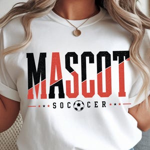 May include: A white t-shirt featuring the word MASCOT in large, distressed black and red block letters. Below the text, the word SOCCER is printed in black with a soccer ball replacing the letter O, flanked by two small red stars on each side.