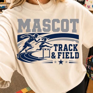 Peut inclure: Un t-shirt couleur crème arborant un motif avec le texte MASCOT en gris et TRACK & FIELD en bleu foncé. Le design comprend une illustration bleu foncé d'un coureur et d'un athlète sautant une haie sur une piste, avec une étoile et des lignes.