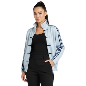 Puede incluir: Chaqueta deportiva azul claro con rayas azul oscuro en las mangas y cuello mandarín. La chaqueta tiene cierre de botones en la parte delantera y se usa con una camiseta negra y pantalones negros. La modelo lleva una coleta alta.