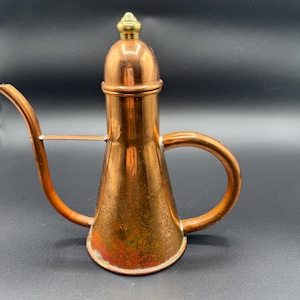 May include: A copper-colored, vintage-style oil pourer with a curved spout and a rounded handle. The pourer has a conical body and a domed lid with a brass finial. The metal has a warm, aged patina, suggesting it is an antique.