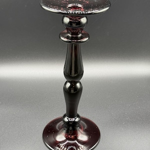 May include: A dark burgundy glass candlestick holder with a round base and a flared top. The stem has a bulbous design with a smooth, glossy finish. The glass has a speckled appearance.