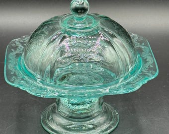 May include: A vintage, teal-colored glass butter dish with a domed lid and pedestal base. The dish features embossed floral and scrollwork patterns. The lid has a small, round handle. The dish is a decorative serving piece.