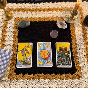 May include: A tarot card reading setup featuring three cards: The Fool, Wheel of Fortune, and King of Pentacles. The cards are on a black surface, surrounded by crystals, a candle, and a gold goblet. The background is a crocheted blanket.