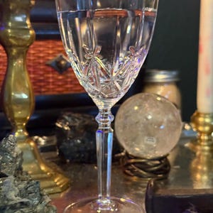 May include: A clear glass goblet featuring a faceted, star-like cut pattern on the bowl. The stem is slender and multi-faceted, leading down to a circular base. The glass is transparent and reflects light, showcasing intricate geometric detailing.