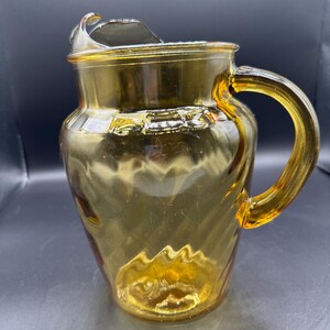 May include: A vintage amber glass pitcher with a handle and a spout. The pitcher has a textured, swirled design and is made of transparent glass. The handle is curved and attached to the side of the pitcher. The pitcher is empty.