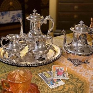 May include: A silver tea set with ornate detailing, including a teapot, creamer, sugar bowl, and serving tray. Two tarot cards and an orange teacup are also visible, set on a patterned tablecloth.