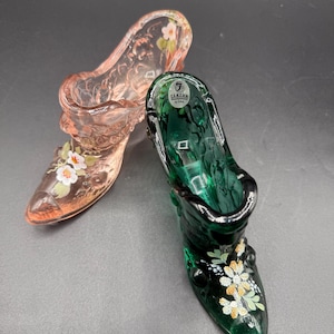 May include: Two glass slipper figurines. One is a translucent pink with white and orange floral accents. The other is a deep green with similar floral details. Both have a classic high-heeled shoe design.