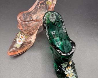 May include: Two glass slipper figurines. One is a translucent pink with white and orange floral accents. The other is a deep green with similar floral details. Both have a classic high-heeled shoe design.