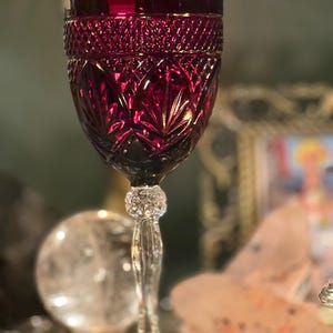 May include: A ruby red wine glass with a detailed, cut-glass design. The bowl features a diamond pattern, and the stem has a decorative, crystal-like element. The glass is set against a blurred background, suggesting a vintage or antique aesthetic.