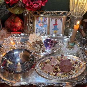 May include: A silver tray holds a collection of spiritual items. A pink crystal ball and rose quartz crystals sit on dried flowers. A lit candle, tarot cards, and red roses complete the display. A small silver bowl and a glass candle holder are also present.