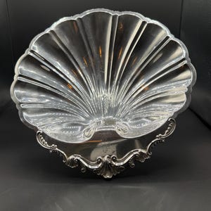 May include: A silver-colored, shell-shaped serving dish. The dish has a fluted design radiating from the center, with a scalloped edge. The base features ornate, scroll-like detailing.