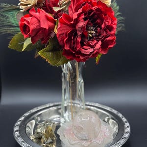 May include: A bouquet of red roses and other flowers in a clear glass vase. The vase sits on a silver-colored tray with a decorative edge. A clear crystal ball rests on a pink and white decorative piece on the tray.