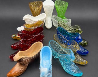 May include: A collection of colorful glass slipper figurines in various shades, including blue, green, red, and yellow. The shoes have a textured surface and are arranged in a semi-circle on a dark background. The collection includes a white vase and other decorative glass pieces.