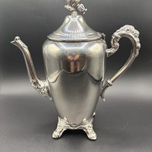 May include: A silver-colored teapot with ornate detailing. The teapot features a curved spout, a decorative handle, and rests on three small feet. The lid is topped with a floral design. The teapot is made of a shiny metal.