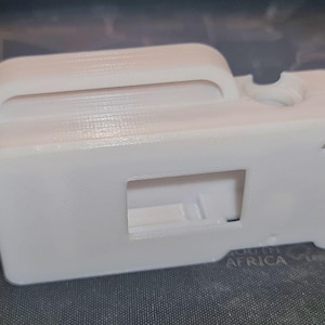 Puede incluir: Un objeto blanco rectangular impreso en 3D con un asa, una abertura rectangular y una pequeña hendidura circular. El objeto tiene una superficie texturizada y parece ser una carcasa para un dispositivo electrónico.