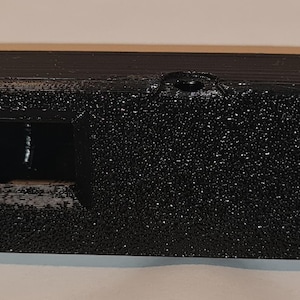 May include: A small, rectangular black object with a textured surface. It has a rectangular opening on one side and a small hole on the top. The object appears to be made of a solid material.