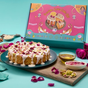 May include: A Persian Love Cake Kit with a frosted cake topped with rose petals and pistachios. The kit includes a box with a cake illustration, lemons, almonds, and rose petals. The cake is on a blue plate, and the kit is on a wooden board.