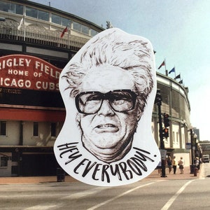 May include: A white sticker with a black and white illustration of a man wearing glasses and the text "Hey Everybody!" The sticker is placed in front of a brick building with the words "Wrigley Field Home of Chicago Cubs" on the side.